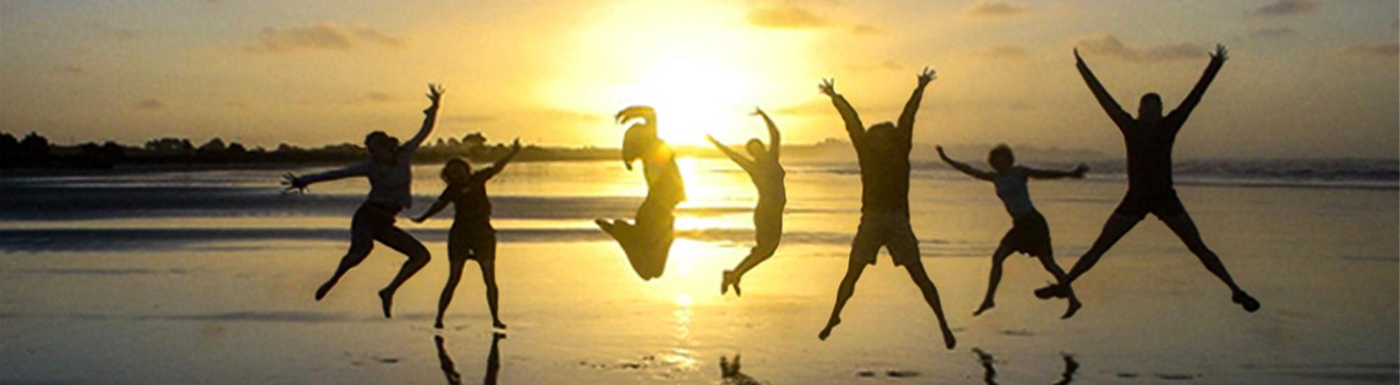 Seven people on beach at sunrise jumping into the air.